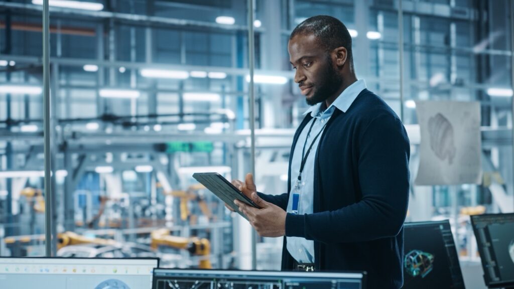 Portrait of Successful Black Male Chief Engineer Using Tablet Computer in Automated Robot Arm Assembly Line Manufacturing High-Tech Electric Vehicles.