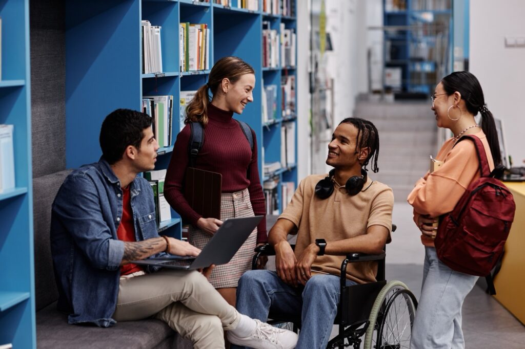 Diverse group of students with young man in wheelchair chatting cheerfully in college library, inclusivity concept