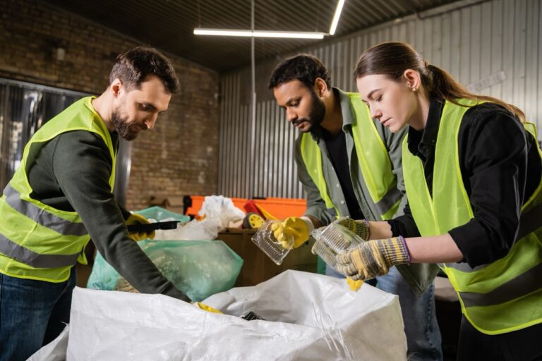 Interracial sorters in protective gloves and safety vests taking plastic containers from sacks while sorting trash together in waste disposal station, garbage sorting and recycling concept