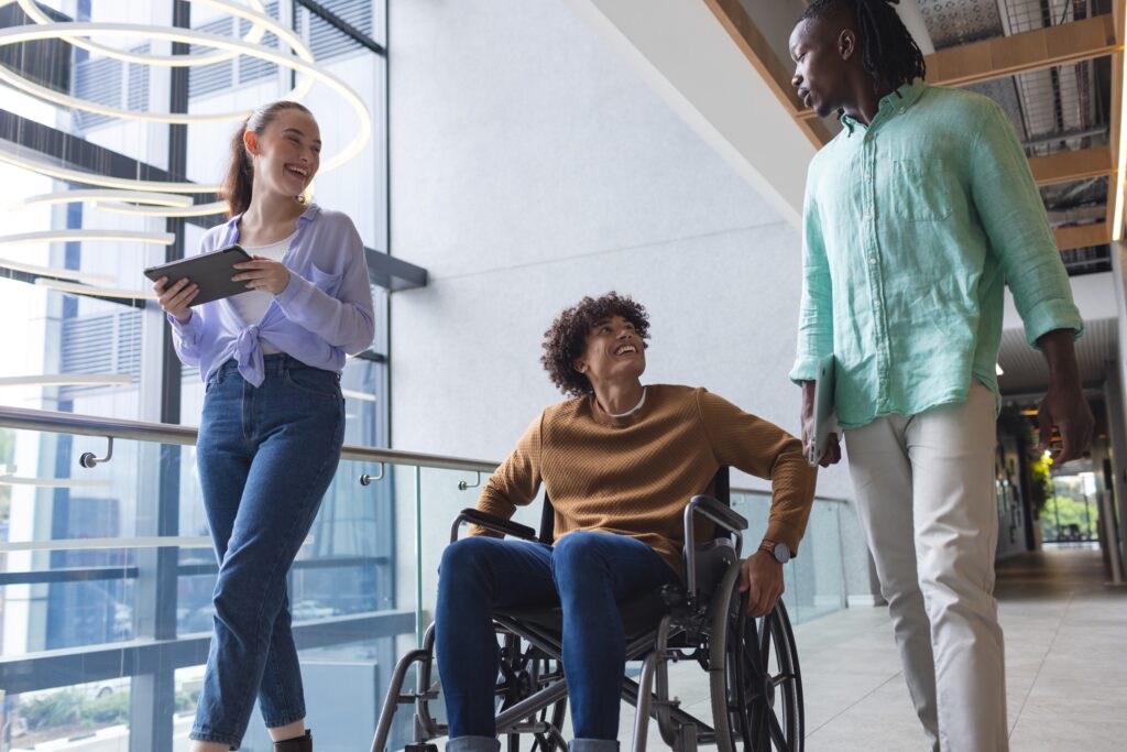 Diverse colleagues walking and talking in bright hallway in a modern business office. African American man wearing green shirt, biracial person in wheelchair, Caucasian woman holding tablet, unaltered