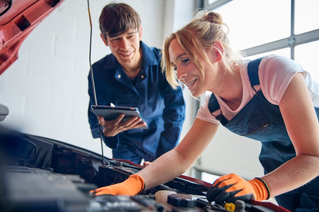 Female Car Auto Mechanic With Trainee Looking Under Bonnet Of Car Using Digital Tablet