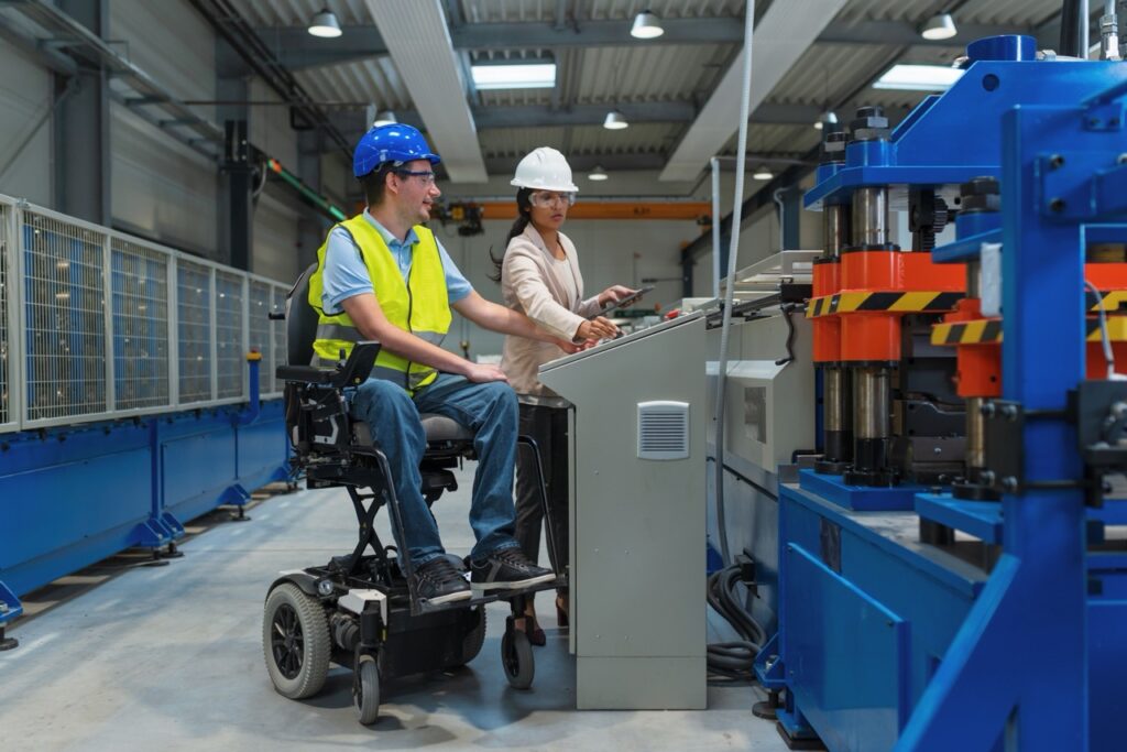 Indian female factory boss training male worker with a disability, in blue helmet and yellow vest, to work on manufacturing machinery.