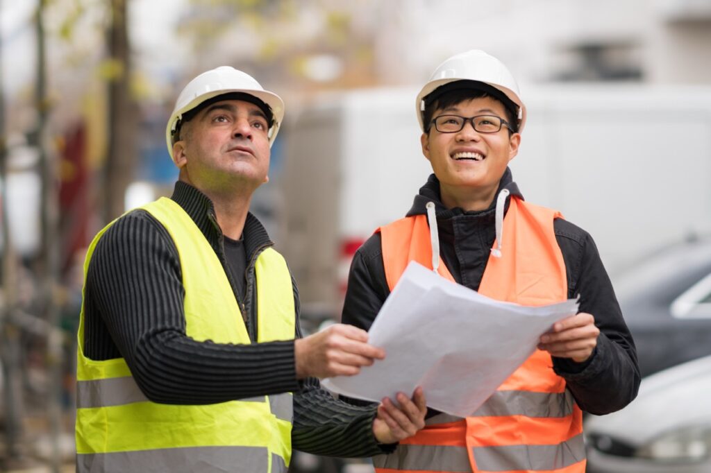 Young Asian apprentice at work on construction site with senior engineer. Outdoors